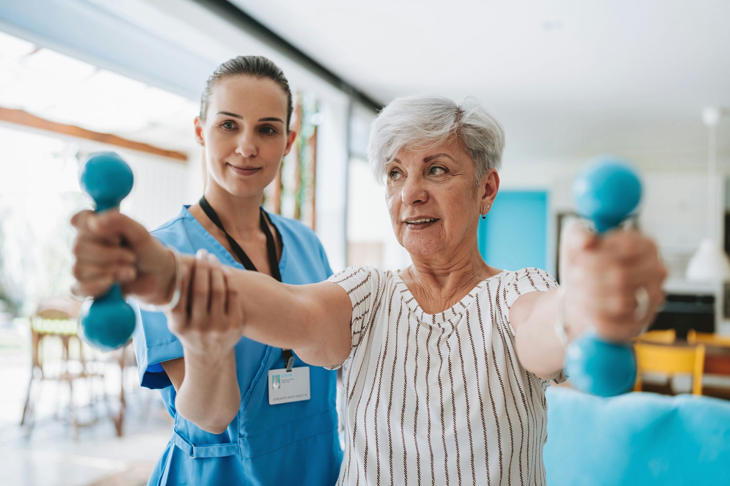 Therapist assisting a senior patient performing front raises with light dumbbells to build shoulder strength.