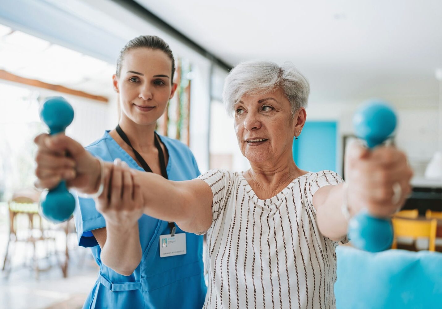 Therapist assisting a senior patient performing front raises with light dumbbells to build shoulder strength.