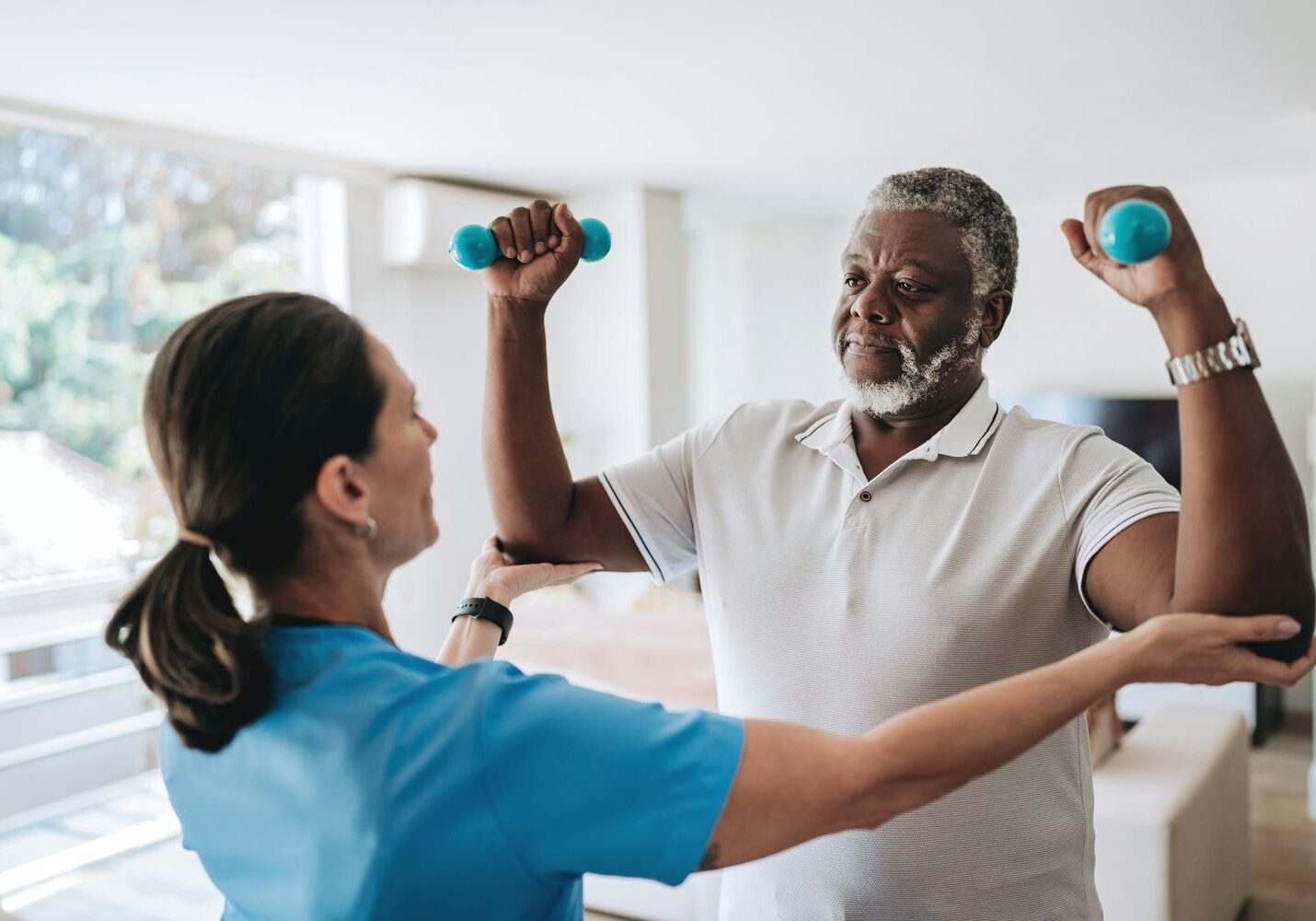 Therapist cueing an older man at home as he lifts light dumbbells to improve shoulder mobility.