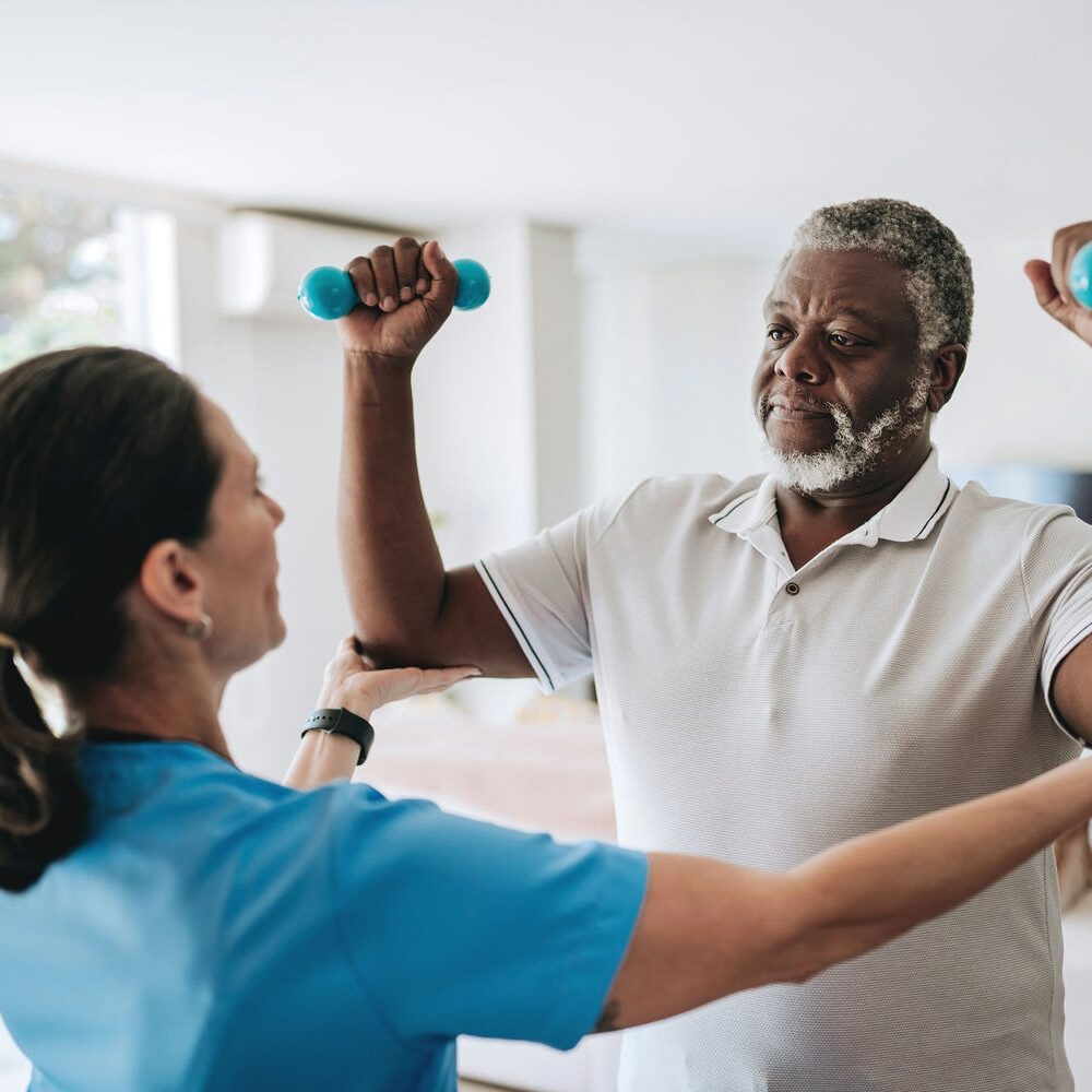 Therapist cueing an older man at home as he lifts light dumbbells to improve shoulder mobility.