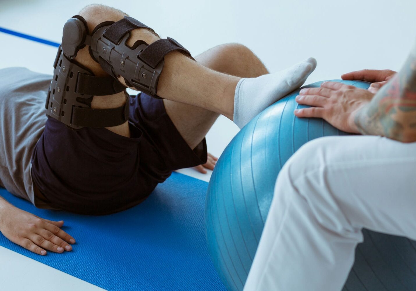 Athlete with a knee brace practicing balance on a therapy ball while the clinician supports the ankle.