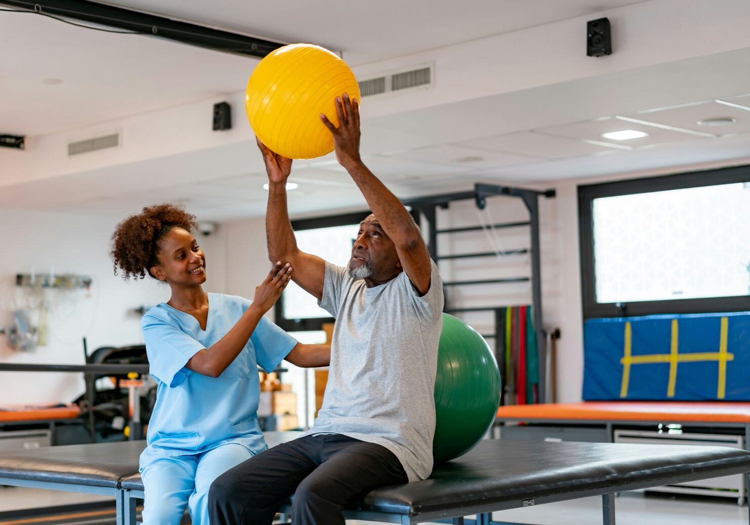 Therapist assisting an adult to lift a yellow therapy ball overhead while seated to build shoulder mobility and core stability.