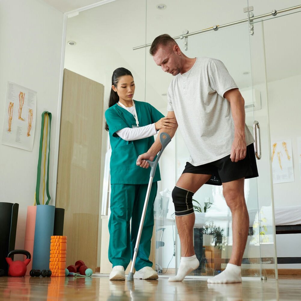 Physiotherapist assisting a patient with a knee brace and cane during clinic rehabilitation.