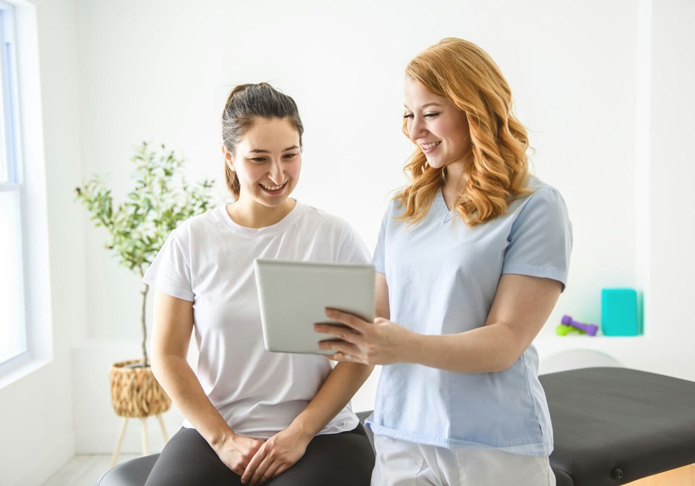 Two women in a physio clinic reviewing exercise progress and home program on a digital tablet.