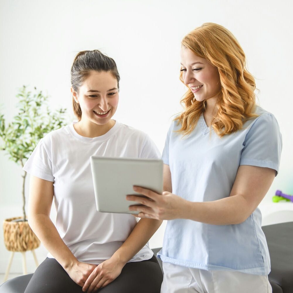 Two women in a physio clinic reviewing exercise progress and home program on a digital tablet.
