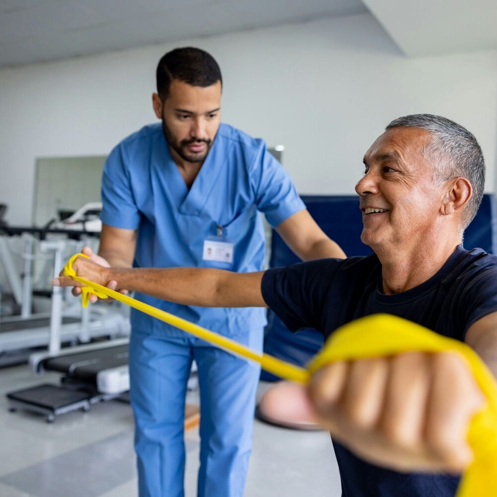 Therapist coaching a patient through shoulder rehab using a resistance band in the gym.