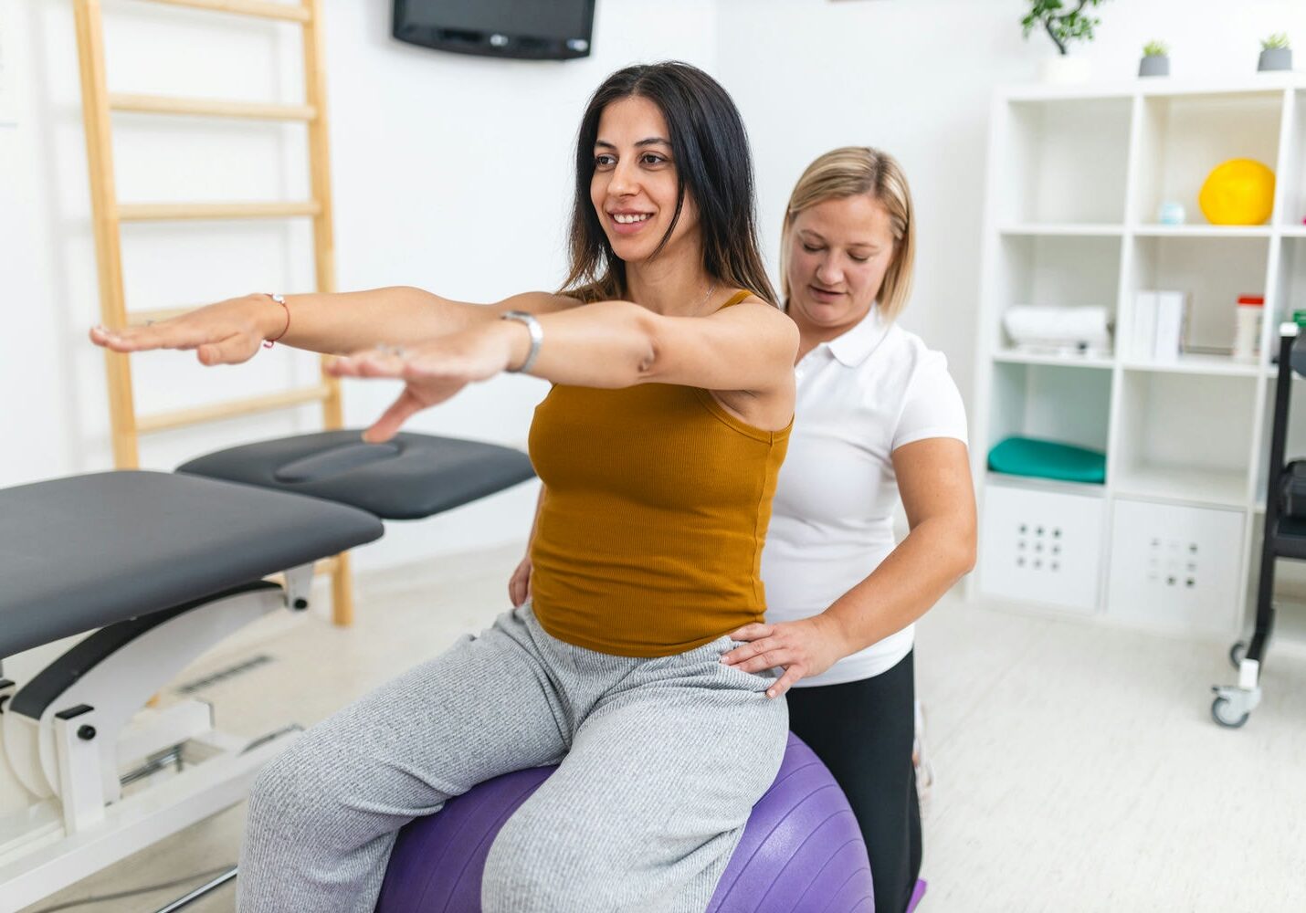Physiotherapist guiding a patient seated on a therapy ball with arms extended for balance and core control.