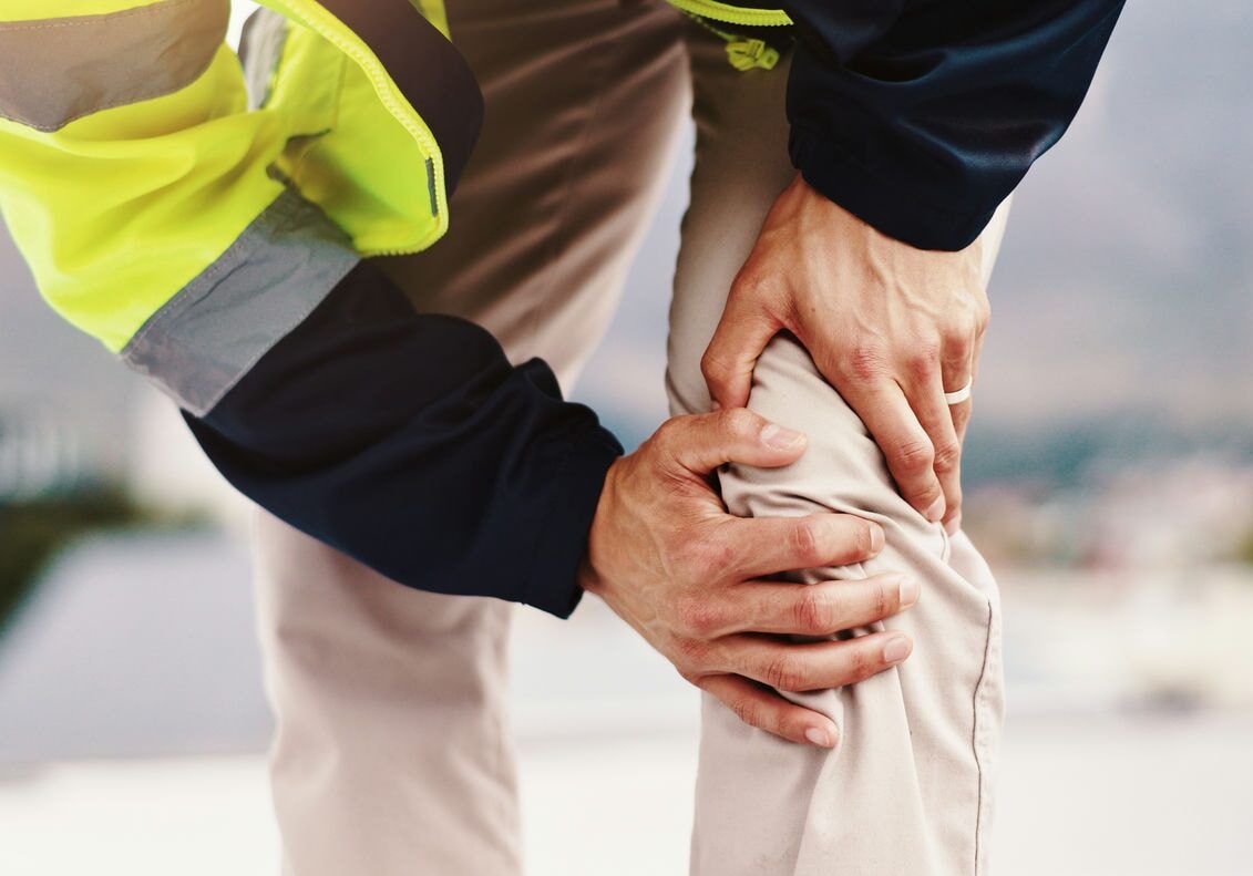 Worker clutching a painful knee outdoors with a helper’s hand providing support, suggesting work-related injury.
