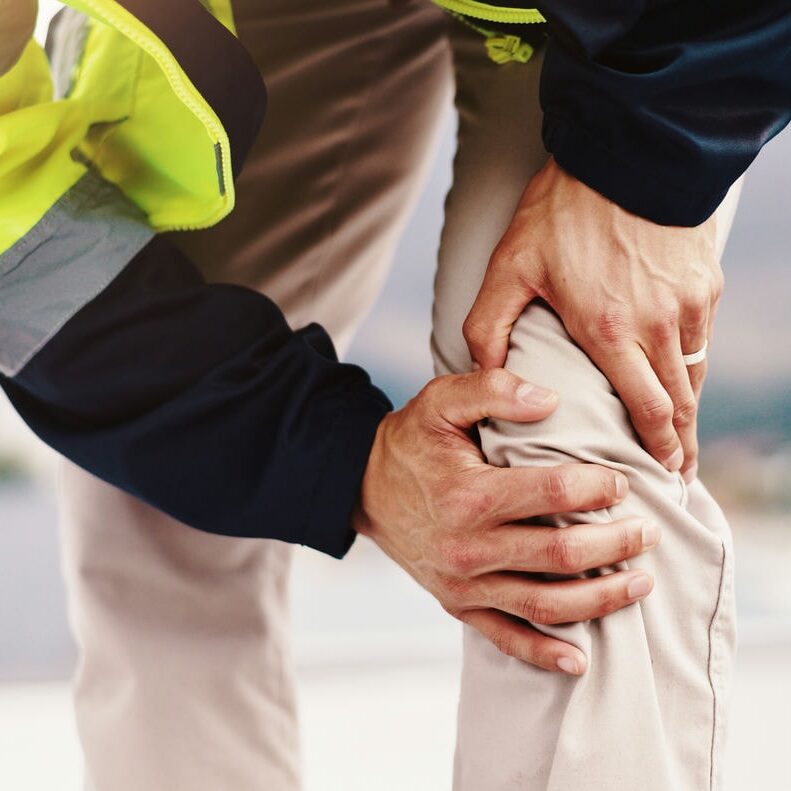 Worker clutching a painful knee outdoors with a helper’s hand providing support, suggesting work-related injury.